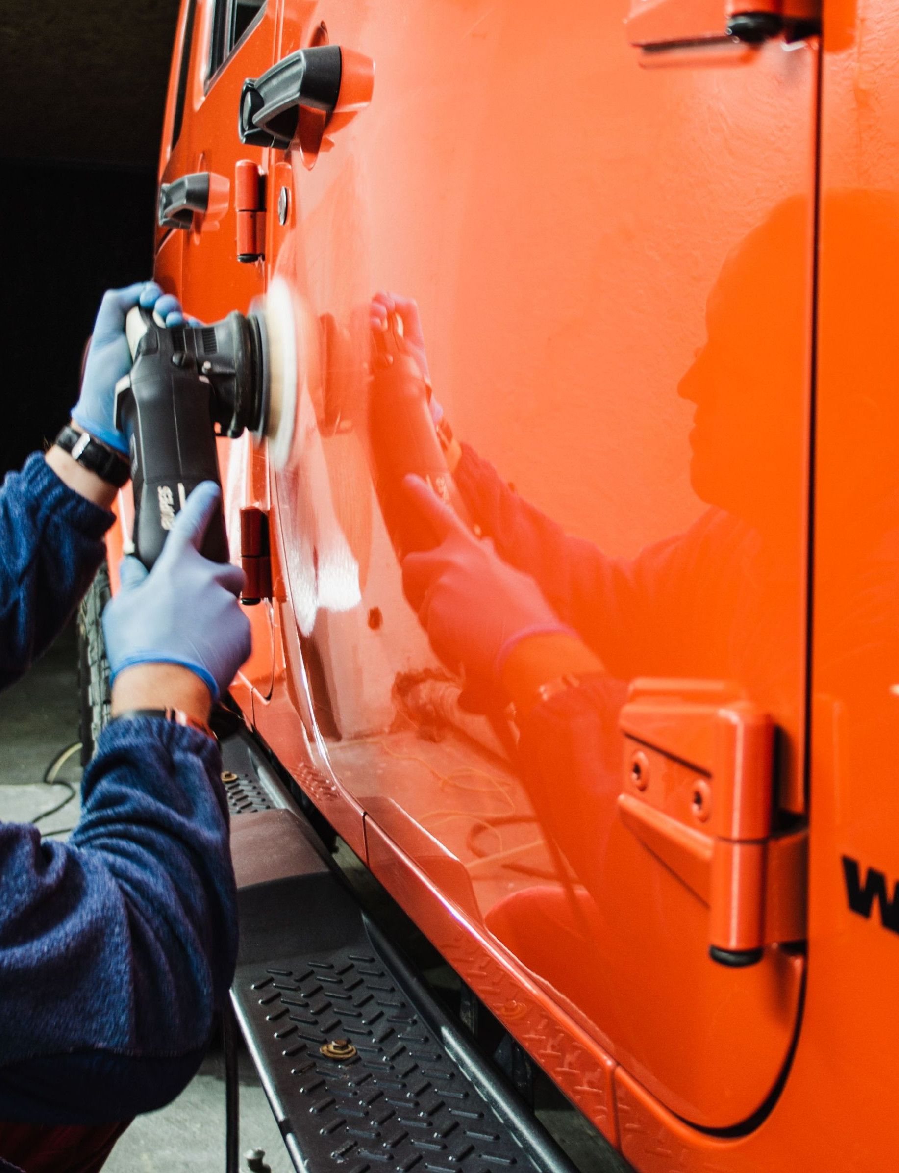 An adult male worker polishes an orange car with a power tool, reflecting dedication to detail.